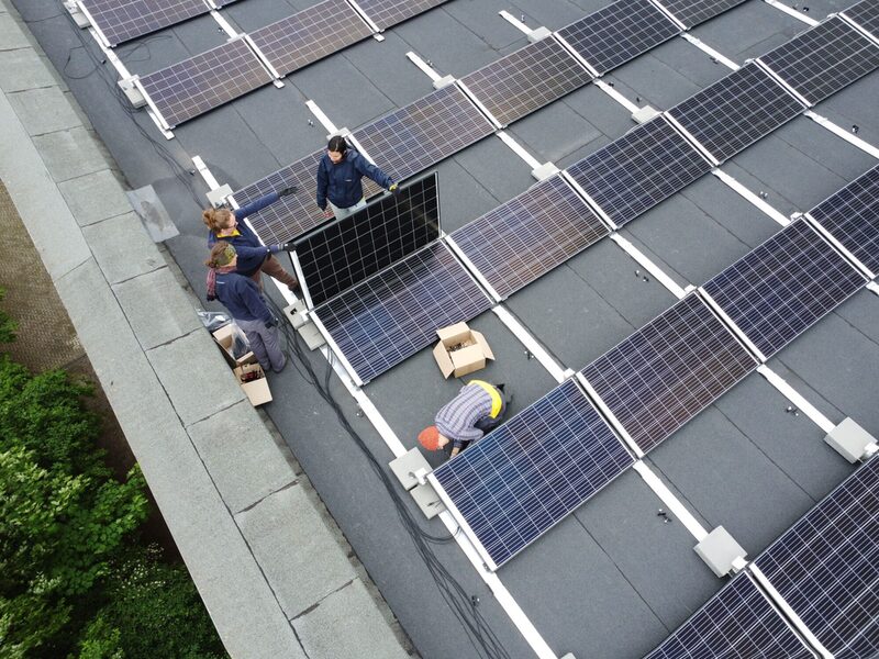 Auf einem grauen Flachdach sind mehrere Photovoltaik-Platten angebracht. Vier junge Erwachsene sind dabei weitere Platten zu befestigen.