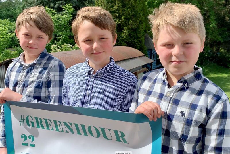 Drei Jungen in blau karierten Hemden stehen nebeneinander und halten ein Banner in der Hand mit der Aufschrift #Greenhour22. Im Hintergrund ein Garten.