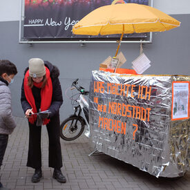 Drei Personen stehen vor einem silberfarbenen Mobil mit orangefarbener Aufschrift.