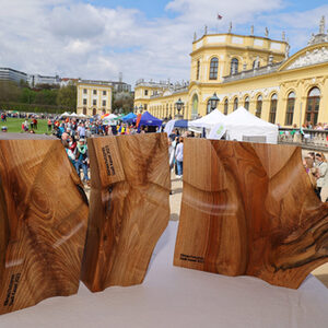 Drei Skulpturen aus Holz, Orangerie und Besuchende im Hintergrund