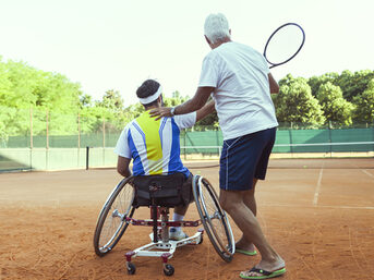 Tennislehrer bringt Mann im Rollstuhl das Tennisspielen bei.