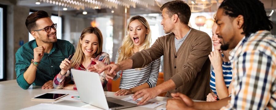 Eine Gruppe junger Menschen steht an einem Arbeitsplatz um einen Laptop herum. Sie zeigen sich erstaunt und glücklich über die Inhalte auf dem Laptop.