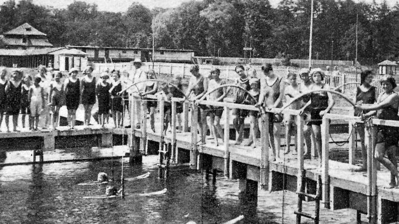 Zahlreiche Menschen im Badeanzug auf einem Steg, im Wasser erste Schwimmerinnen.