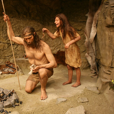 Eine lebensechte Nachbildung eines Mädchens hält eine Maus in den Nacken des vor ihr knienden Vaters in einer Steinzeit-Szene.
