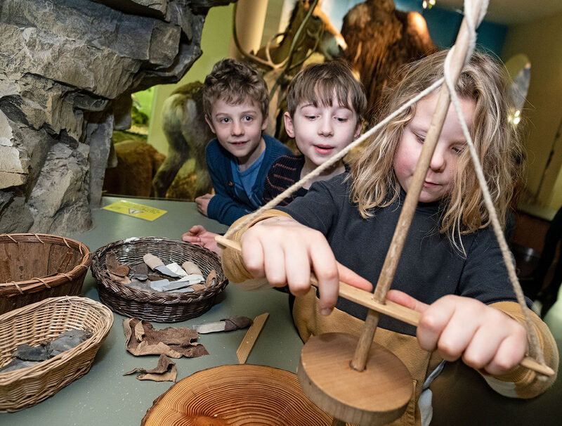 Kinder bohren mit einem Steinzeitbohrer aus Holz auf einer Scheibe.
