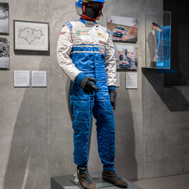 An einer Schaufensterpuppe ist der Anzug und der Helm eines Rennfahrers ausgestellt, sowie ein Pokal und Bilder von einem Rennauto.